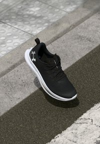 Zapatilla deportiva negra con parte superior de malla, suela exterior blanca y cordones negros. Cuenta con un logo de Under Armour en el lateral y un collar acolchado.