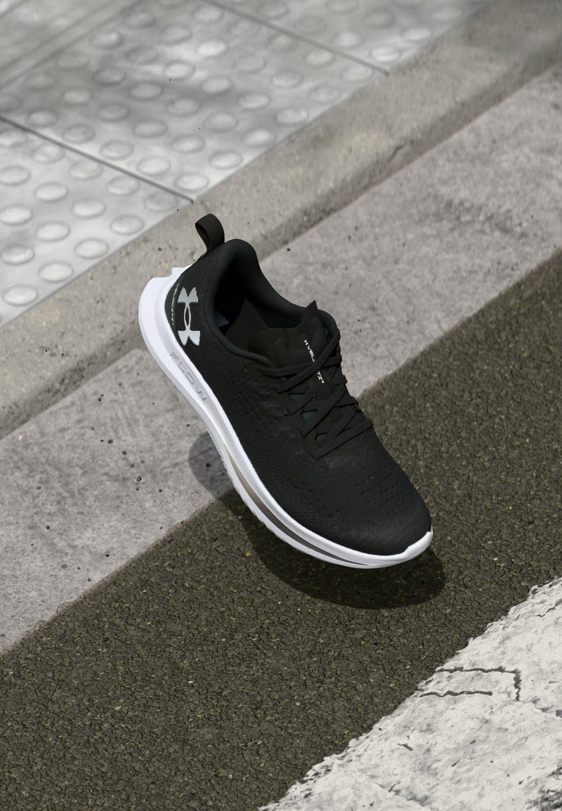 Zapatilla deportiva negra con parte superior de malla, suela exterior blanca y cordones negros. Cuenta con un logo de Under Armour en el lateral y un collar acolchado.