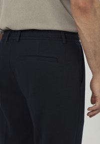 Marineblauwe trainingsbroek met een gladde textuur, elastische tailleband en achterzak met paspel. Geplaatst naast een lichtbeige shirt.