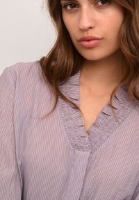 Blusa viola chiaro con strisce verticali, caratterizzata da un collo a V arricciato e una consistenza morbida. Il tessuto sembra leggero e fluido.