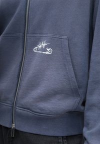 Tengerészkék cipzáras pulóver elülső zsebbel; fehér hímzett biztonsági tű és csillagok mintával; puha, bolyhos tapintású anyagból.