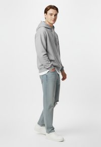 Felpa grigia con tasca frontale e cordino, abbinata a jeans azzurri chiari con orlo strappato e sneakers bianche.