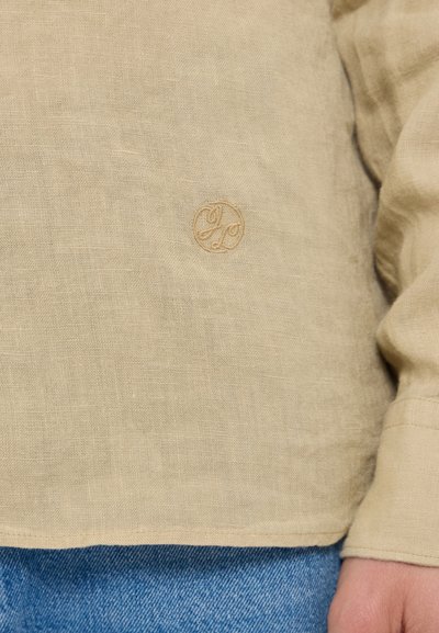Beige lněná košile s dlouhým rukávem a jemně vyšívaným logem na hrudi. Poskytuje volný střih a texturovanou, přirozenou tkaninu.