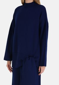 Maglione lavorato a maglia blu navy con collo alto, vestibilità oversize e dettaglio annodato in vita. Texture morbida e maniche lunghe.