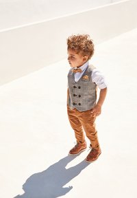 Child wearing a gray plaid vest, white shirt, and orange bow tie, paired with brown trousers and brown shoes. Light source casts a shadow.