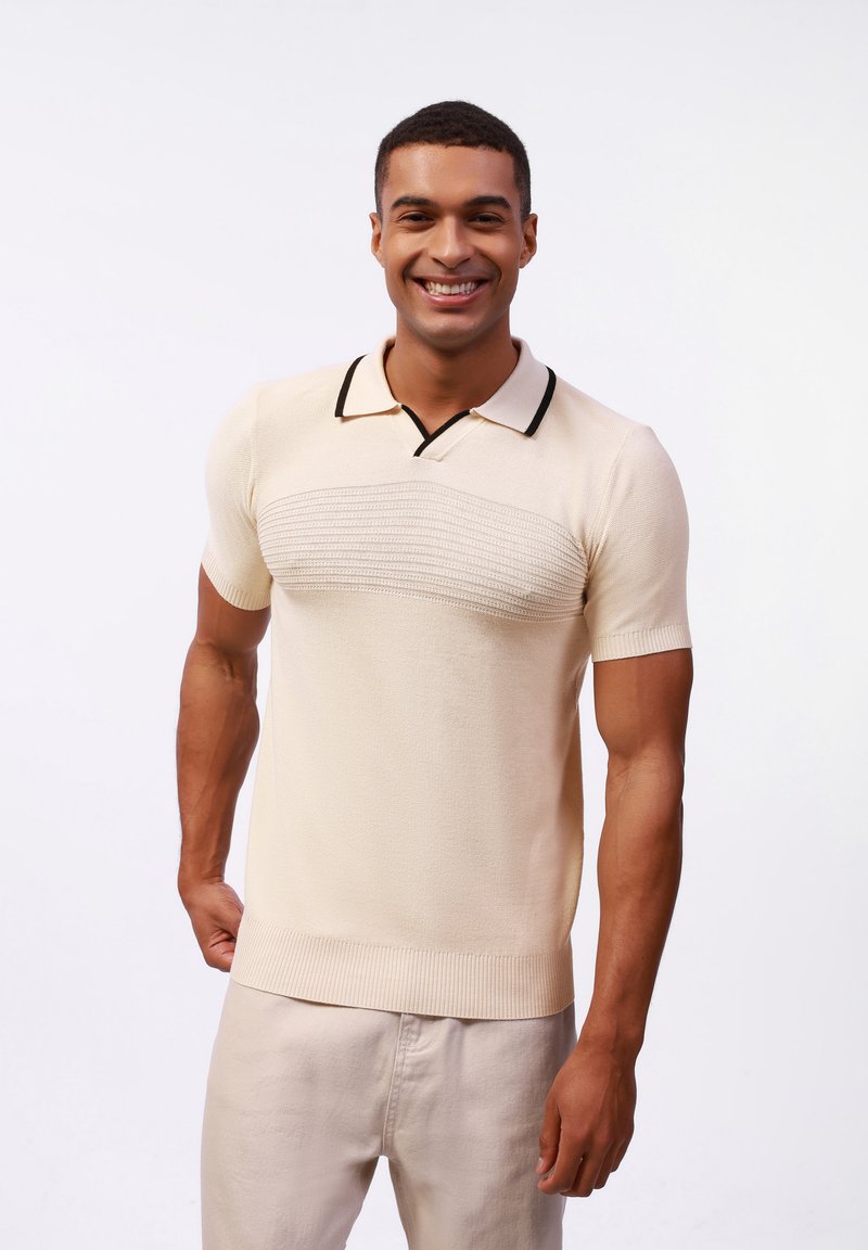 Beige polo triko s krátkým rukávem, se strukturovaným horizontálním pruhem přes hruď, černým lemem límečku a žebrovanými okraji spodního lemu a rukávů.