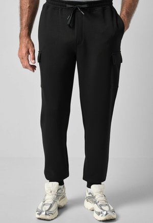Mand iført sorte cargo joggingbukser med snøre i taljen og hvide chunky sneakers, stående mod en ensfarvet baggrund.