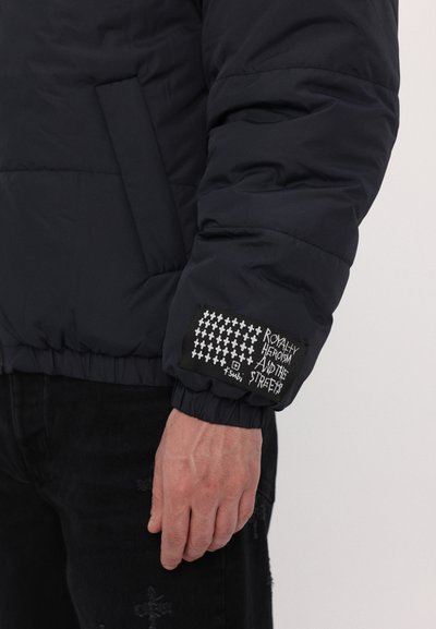 Veste en duvet bleu marine avec texture matelassée, poignets élastiques et un patch en tissu noir sur la manche arborant du texte blanc et un motif contrastant.