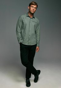 Green checkered button-up shirt with long sleeves, paired with black pants and black shoes, set against a neutral background.
