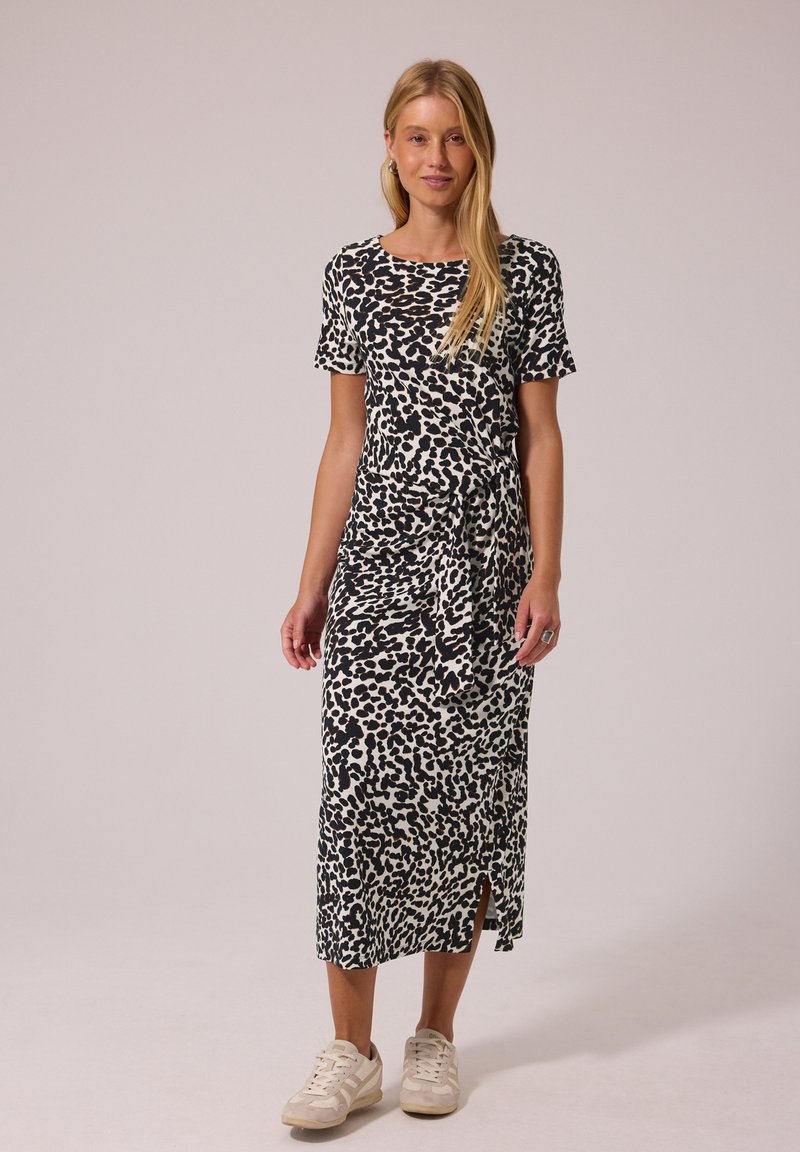 Femme portant une robe midi noire et blanche à pois, manches courtes et baskets, debout devant un fond clair uni.