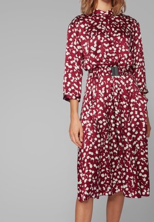 Femme portant une robe bordeaux mi-longue avec des pois blancs abstraits, manches trois-quarts, et une ceinture noire cintrée à la taille.