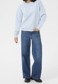Felpa azzurra chiara con stampa "PARIS", abbinata a jeans a gamba larga blu scuro. Entrambi hanno una vestibilità casual e una texture liscia. Scarpe nere completano il look.