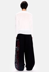 Balti ilgomis rankovėmis marškiniai su permatomu tekstūra, derinami su laisvomis juodomis kelnėmis, kurių šonuose yra raudoni ir balti abstraktūs raštai.