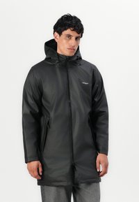 Parka nera impermeabile con cappuccio, chiusura frontale con zip, tasche laterali e finitura opaca. Dettagli testurizzati su polsini e orlo.