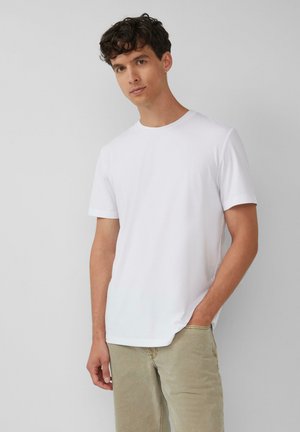 Weißes Baumwoll-T-Shirt mit Rundhalsausschnitt und kurzen Ärmeln, das eine glatte Textur und eine lockere Passform aufweist, kombiniert mit hellbeigen Hosen.