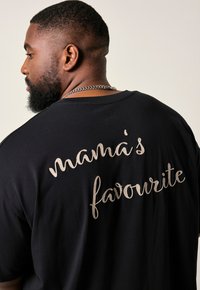 Sort bomulds T-shirt med stor broderet tekst på ryggen, der siger "mamas yndling" i beige. Løs pasform.
