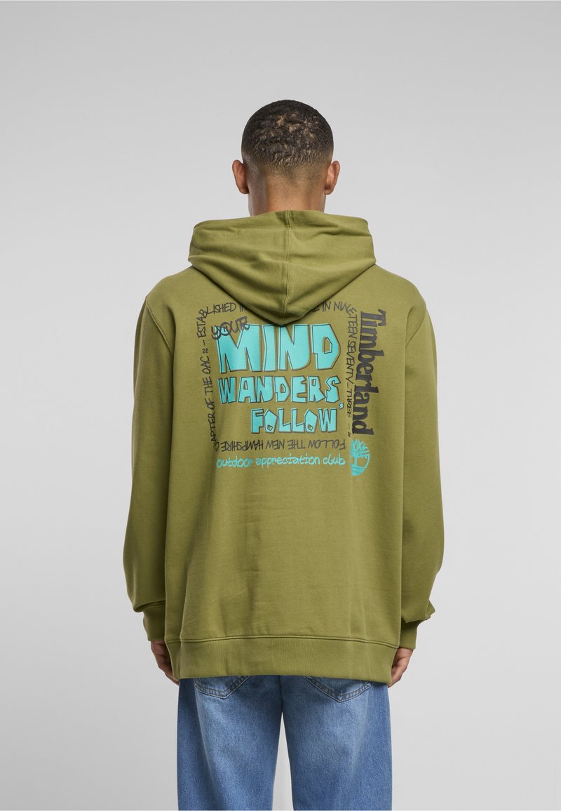 Férfi olíva zöld Timberland kapucnis pulóverben, kék felirattal: "Your mind wanders follow" és farmerben, háttal a szürke háttér előtt.