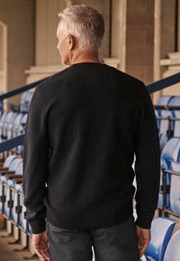Juodas megztinis su ilgomis rankovėmis, apvaliu kaklu ir raukšlėtais rankogaliais. Dėvimas su tamsiomis kelnėmis, matomas iš nugaros stadiono aplinkoje.