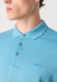 Svetlo modra polo majica iz mešanice bombaža, z klasičnim ovratnikom, tri gumbi in vezenim logotipom "BOSS" na prsih.