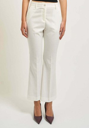 Donna che indossa pantaloni bianchi su misura a gamba dritta e scarpe con tacco alto scure e punta affilata, in piedi su uno sfondo neutro.