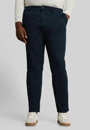 Homme portant un pantalon bleu marine foncé, des baskets blanches et une chemise blanche à manches longues avec de fines rayures grises, debout, une main dans la poche.