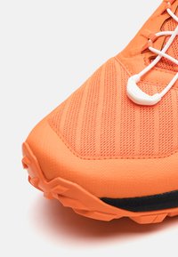 Orange atletisk sko med texturerad ovandel, förstärkt tåhätta och kontrasterande svart sula med multidirektionella greppklumpar.