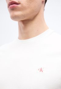 Νέος άνδρας που φοράει απλό λευκό μπλουζάκι με μικρό κόκκινο λογότυπο Calvin Klein στο στήθος, με λευκό φόντο.