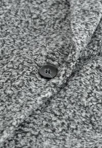 Tejido gris texturizado con una apariencia peluda. Presenta un solo botón negro con dos agujeros. La tela muestra un patrón de color gris mezclado.