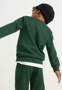 Grøn sweatshirt lavet af blødt stof, med ribbede kanter ved hals og ærmer. Matchende grønne bukser og en sort strikhue.