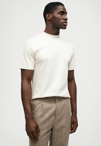 Tricou crem cu mânecă scurtă și decolteu rotund, asortat cu pantaloni texturați maro. Materialul pare neted și ușor.