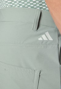 Gaiši zaļi sporta šorti, izgatavoti no mīksta auduma, ar aizmugurējo kabatu un baltām šuvēm, ar nelielu baltu Adidas logotipu.