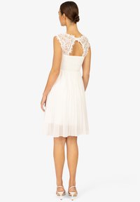 Robe blanche avec un corsage en dentelle aux motifs floraux, une découpe au dos et une jupe plissée. Texture lisse et design mi-long.