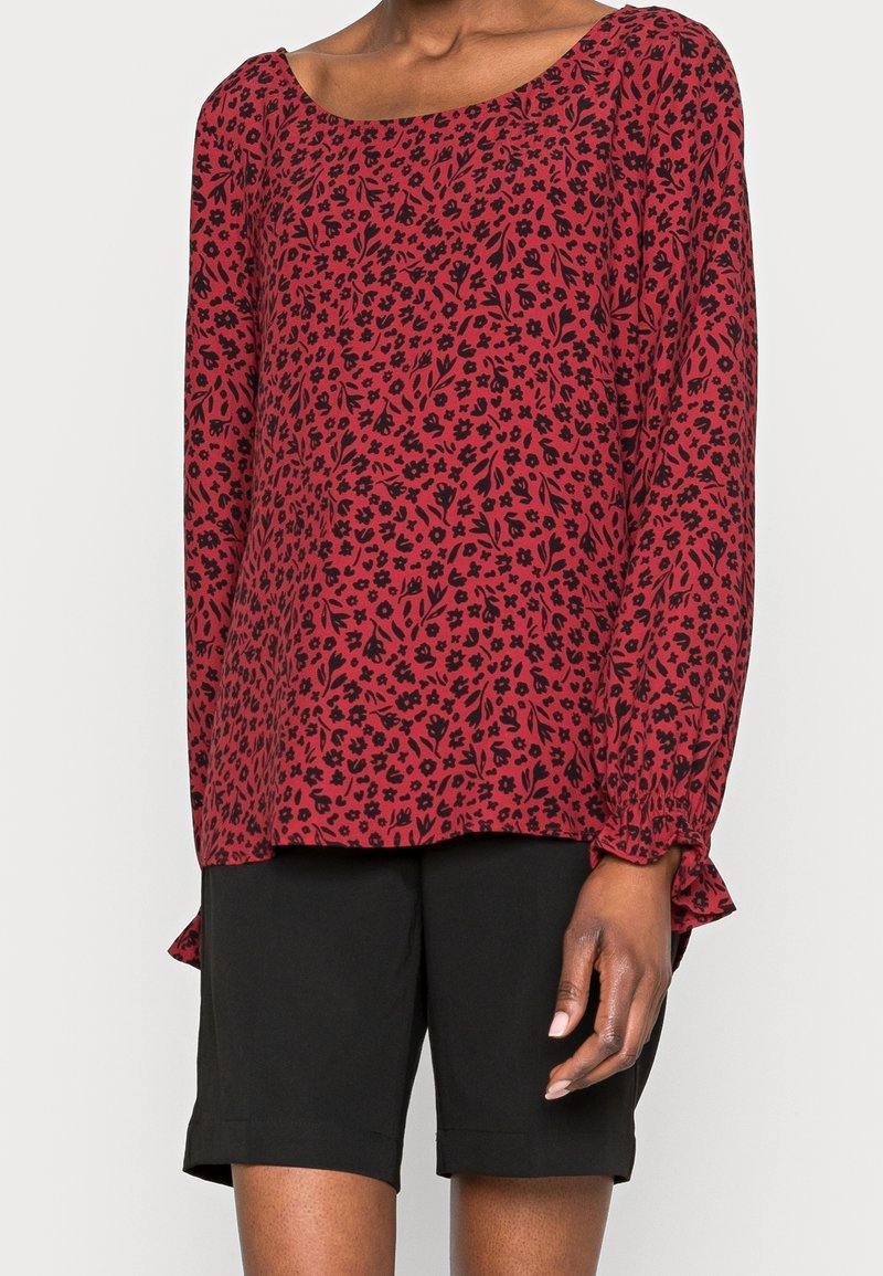 Femme portant un chemisier à manches longues rouge avec un imprimé floral noir et un short noir arrivant aux genoux, debout devant un fond clair.