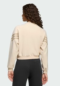 Žena s kučeravými vlasmi, oblečená v béžovom topu s dlhým rukávom a tromi pruhmi na rukávoch, a čiernych nohaviciach, stojí chrbtom k jednoliatemu pozadiu.