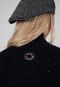 Pull texturé bleu marine avec col côtelé et détail logo brodé beige au dos. Casquette à chevrons grise partiellement visible.