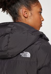 Crna puffer jakna s kapuljačom, s qiltanom teksturom i bijelim logom "The North Face". Vidljivo šivanje i moderan silueta.