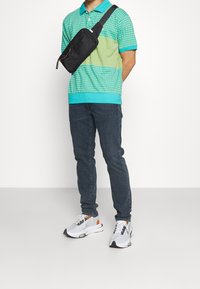 Person som bär en turkos- och gulmönstrad pikétröja, mörka jeans, vita sneakers och en svart axelväska framför en helt vit bakgrund.