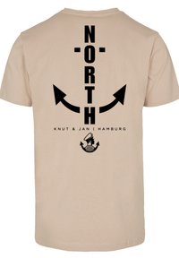 Beige t-shirt featuring black text "NORTH" with arrows and a logo of a sailor's face and waves at the bottom. Soft fabric, casual design.