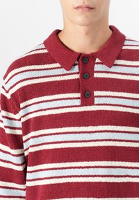 Strikket polo skjorte med bordeaux, kremfargede og lyseblå horisontale striper, med en strukturert finish og tre svarte knapper ved kragen.