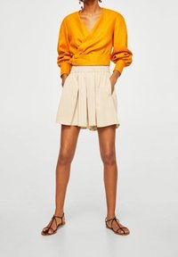 Femme portant un haut orange à manches longues avec manches bouffantes, un short beige taille haute plissé, et des sandales plates noires à lanières.
