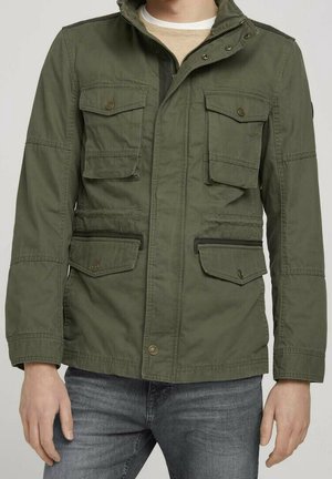 Mann trägt eine olivgrüne Utility-Jacke mit vier Klappentaschen und Knopfdetails, kombiniert mit blauen Jeans und einem beigen Hemd darunter.