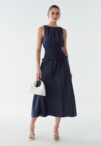 Abito senza maniche blu navy con vita arricciata e dettagli arricciati; abbinato a una borsa bianca con manico ad anello dorato.