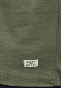 Oliven grøn bomull T-skjorte med en strukturert overflate, med en hvit etikett sydd i sømmen med "BLEND 1993" og "BUILT IN BLUE."