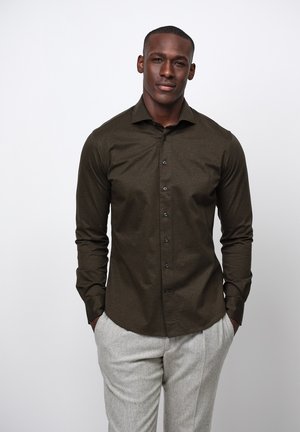 Camisa de botón en verde oliva oscuro con un corte ajustado, con botones negros y una textura suave. Combinada con pantalones gris claro.