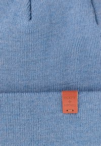 Blauwe gebreide beanie met een omgeslagen rand, voorzien van een tan leren merkpatch met het label "BICKLEY + MITCHELL." Gestructureerde stof met fijne steken.
