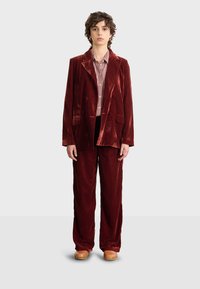 Szkarłatny aksamitny garnitur z podwójnym zapięciem i dopasowanymi spodniami w szerokim kroju, uzupełniony o koszulę w kratkę i drewniane chodaki.