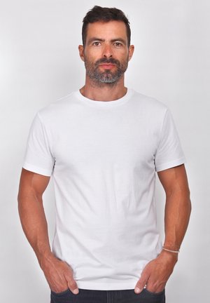 Balta īss piedurkņu T-krekls no kokvilnas ar apaļu kakla izgriezumu un gludu tekstūru. To valkā bārddzīlis, rokas ielikts kabatās.