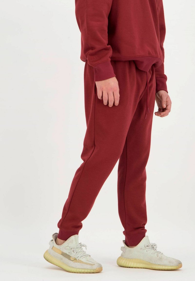Bordeaux-rotes Sweatshirt und passende Jogginghose aus gebürstetem Stoff, mit verstellbarem Bund, gerippten Bündchen und kontrastierenden Nähten. Getragen mit hellen Sneakers.