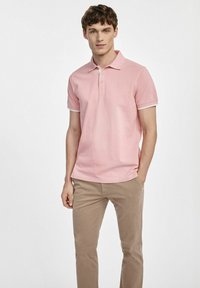 Jasnoróżowa bawełniana koszulka polo z kołnierzykiem, dwoma guzikami i kontrastującymi białymi mankietami, w połączeniu z beżowymi chinosami, niewielka widoczność marki.