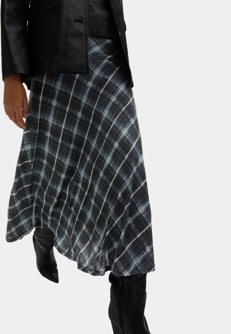 Vrouw die een zwart leren jas draagt, een donkere asymmetrische geruite rok en zwarte laarzen met spitse neus en hakken, lopend naar voren.